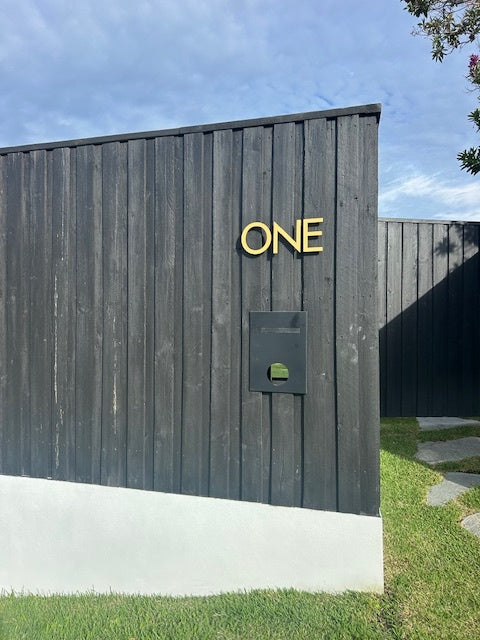 The word ONE in matt gold capital letters on a black vertical-board fence, above a black letterbox