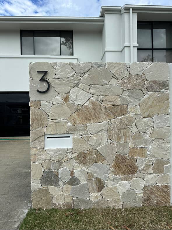 A large matt black 3 on a stone-clad letterbox pier with a white letter slot, white two-storey house behind